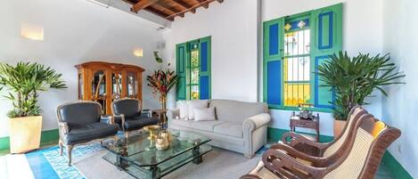 A spacious living room with a glass coffee table, a sofa, and two armchairs. There are potted plants and a wooden cabinet.