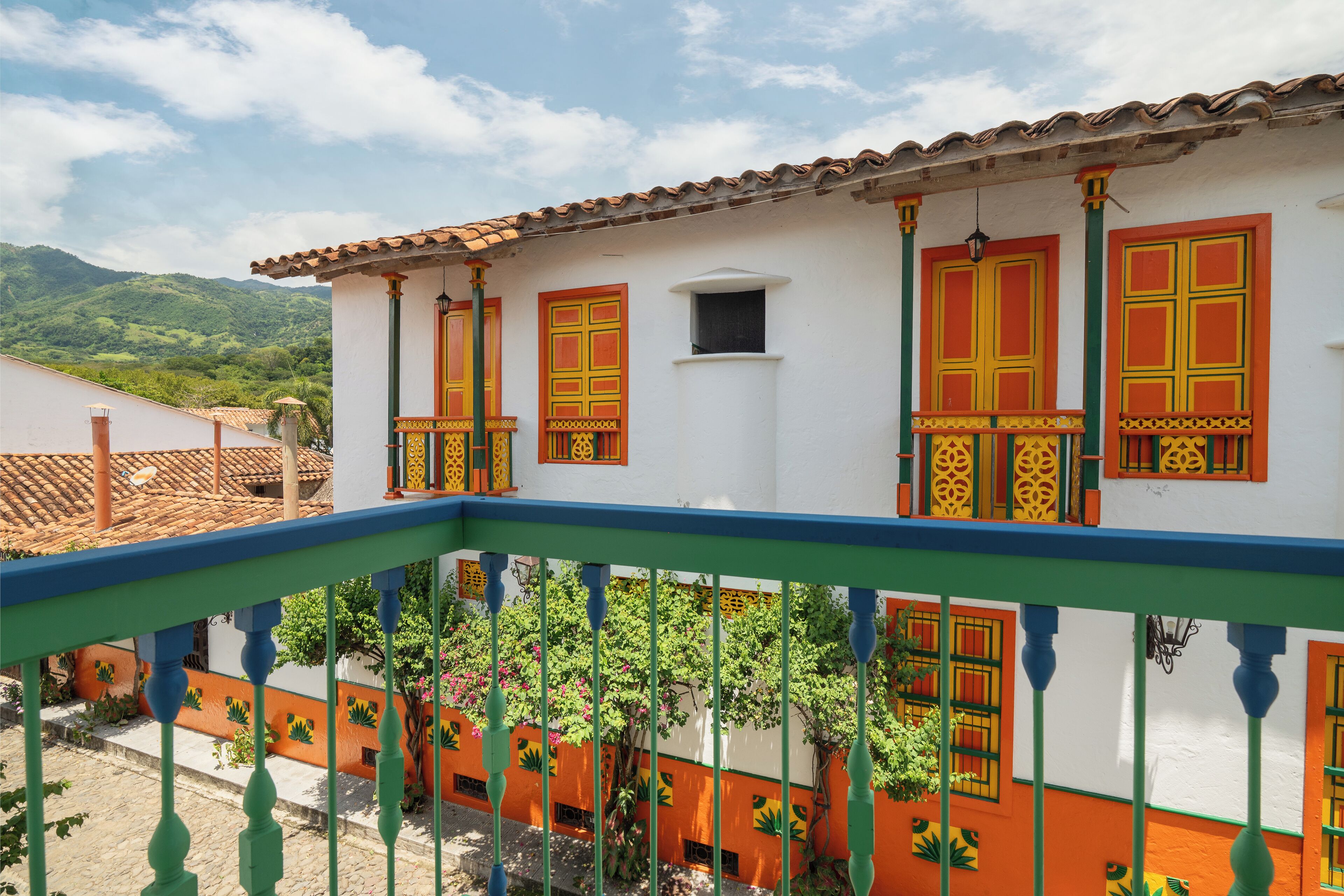 Ein weißes Gebäude mit orange- und grünfarbigen Akzenten, ein Balkon mit blauen Geländern und eine gebirgige Landschaft im Hintergrund.