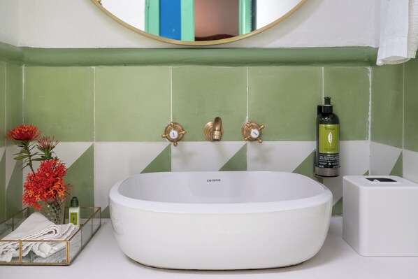 A bathroom with a white sink, green and white tiles, a mirror, a bottle of shampoo, and a vase with flowers.