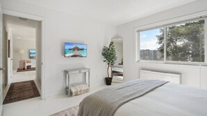 Ein modernes Schlafzimmer mit einem großen Bett, einem an der Wand montierten Fernseher, einem Blumentopf und Blick auf die Stadt durch große Fenster.