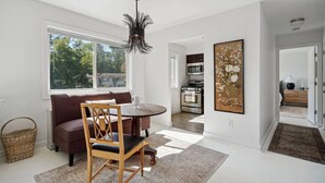 Ein modernes Wohnzimmer mit Sofa, Esstisch und Stühlen, mit einer Küche im Hintergrund und einem großen Fenster, das natürliches Licht hereinlässt.