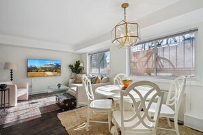 A modern living room with a white sofa, a glass coffee table, and a dining table with white chairs. There is a large window with a view of a building outside. - Executive King Suite / 5 min to DCA Pentagon HQ2 / Free Parking (Arlington)