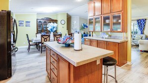 A kitchen with wooden cabinets, a central island with stools, a dining area with a table and chairs, and a living room with a couch and curtains. - Lake Livingston Home w/ Dock & Deck! (Huntsville)