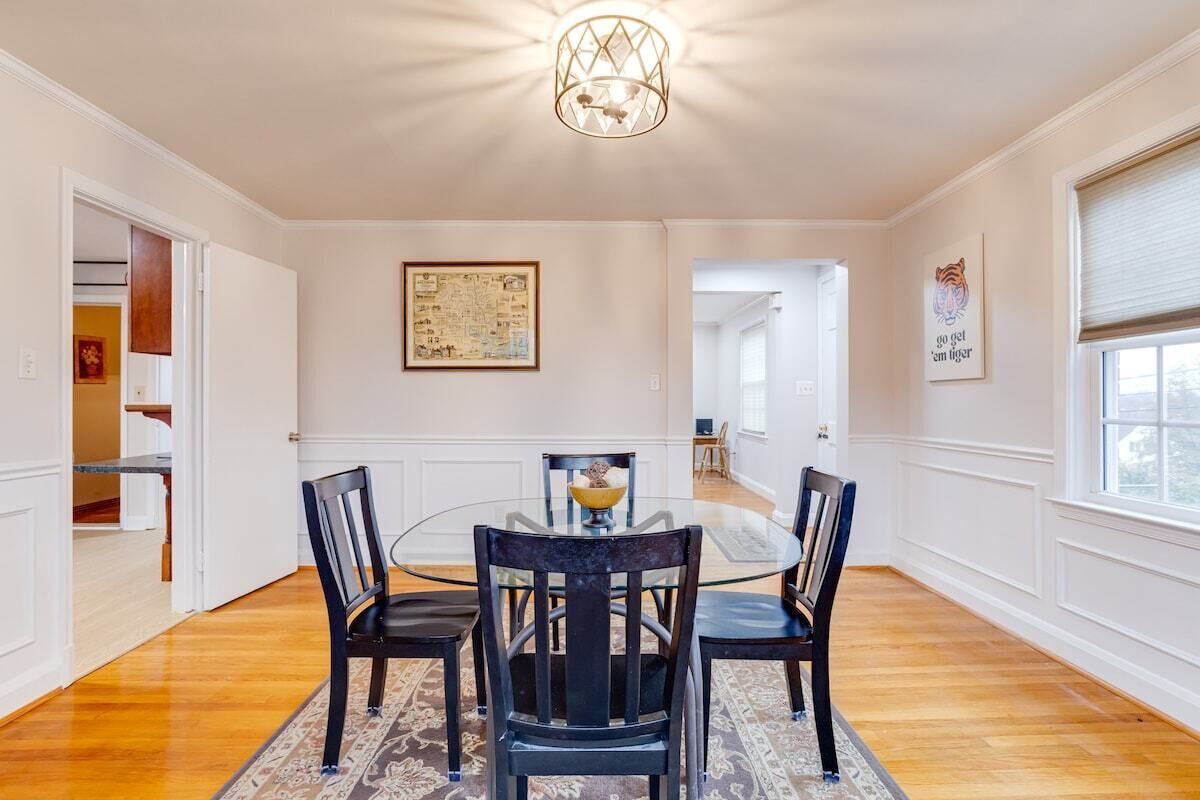 A dining area with a round glass table, four chairs, a chandelier, a map on the wall, and a window with blinds.