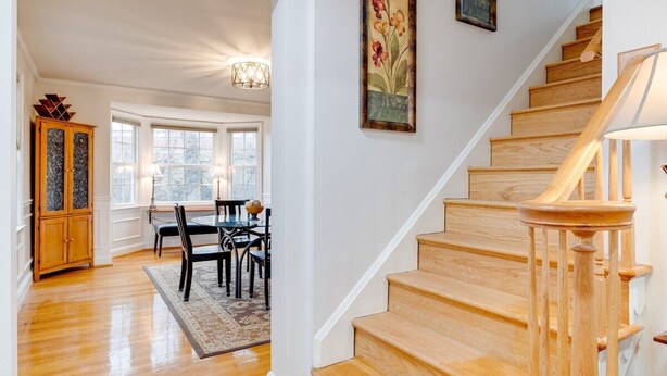 A staircase with wooden steps and a handrail, leading to a dining area with a table and chairs, and a view of the outside through large windows. - Welcome To Tigertown! 4 Bd House (Towson)