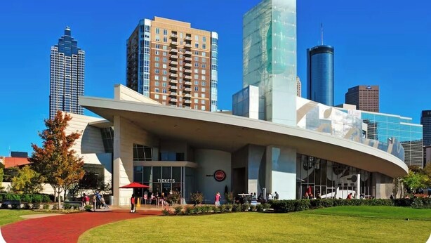 Um edifício moderno com telhado curvo e uma placa com a palavra 'Tickets', rodeado por um gramado bem cuidado e outros edifícios altos ao fundo.