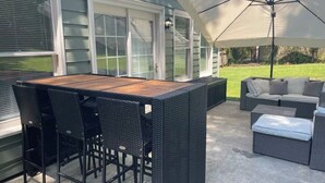 Um pátio externo com mesa de madeira e cadeiras de vime preto, um sofá cinza e um grande guarda-sol.