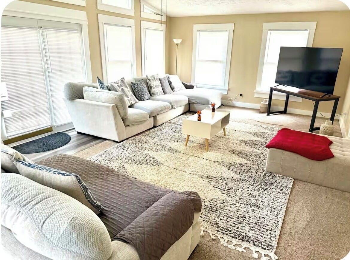Ein Wohnzimmer mit einem Ecksofa, einem Couchtisch, einem Fernseher auf einem Schrank und einem großen Fenster mit Jalousien.