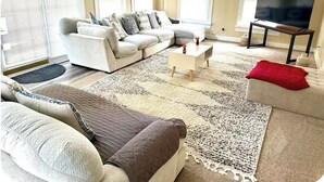 Sala de estar com sofá seccional, mesa de centro, televisão em um suporte e uma janela grande com persianas.