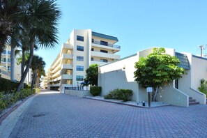 A paved walkway leading to a modern building with multiple floors and balconies, surrounded by palm trees and a clear sky.