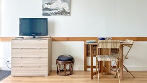 A room with a television on a wooden cabinet, a small stool, a wooden chair with a cushion, and a painting of a lighthouse on the wall. - Large studio with breathtaking views of the Baie des Sables (Les Sables-d´Olonne)
