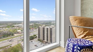 A room with a large window offering a city view, a brown cushioned chair, and a blue patterned stool. - 20th FL 1BR | Heated Pool | Gym | Bar | Balcony (Austin)