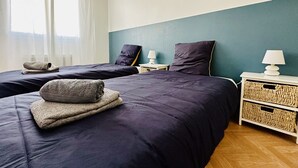 Habitación de hotel con dos camas, ventana con cortinas translúcidas, pared de acento color verde azulado y una mesita de noche con lámpara y cesta tejida.