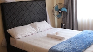 Un lit bien fait, avec une tête de lit matelassée, une serviette enroulée et un ventilateur sur pied.