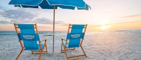 Duas cadeiras de praia azuis com a marca "Margaritaville Beach Cottage Resort", sob um guarda-sol azul correspondente, numa praia arenosa com o oceano ao fundo.