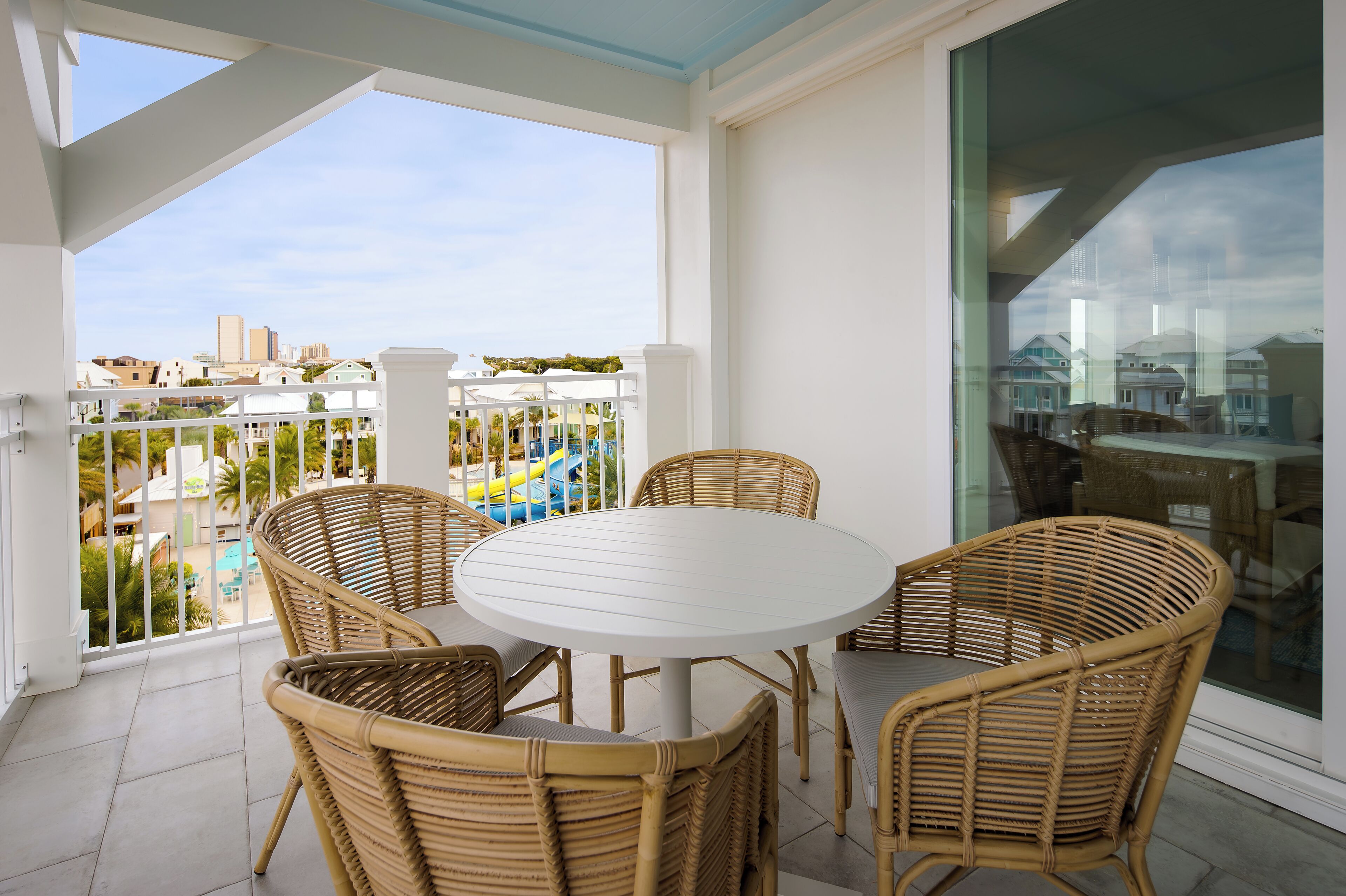 A balcony with wicker chairs and a white table, overlooking a pool and buildings.