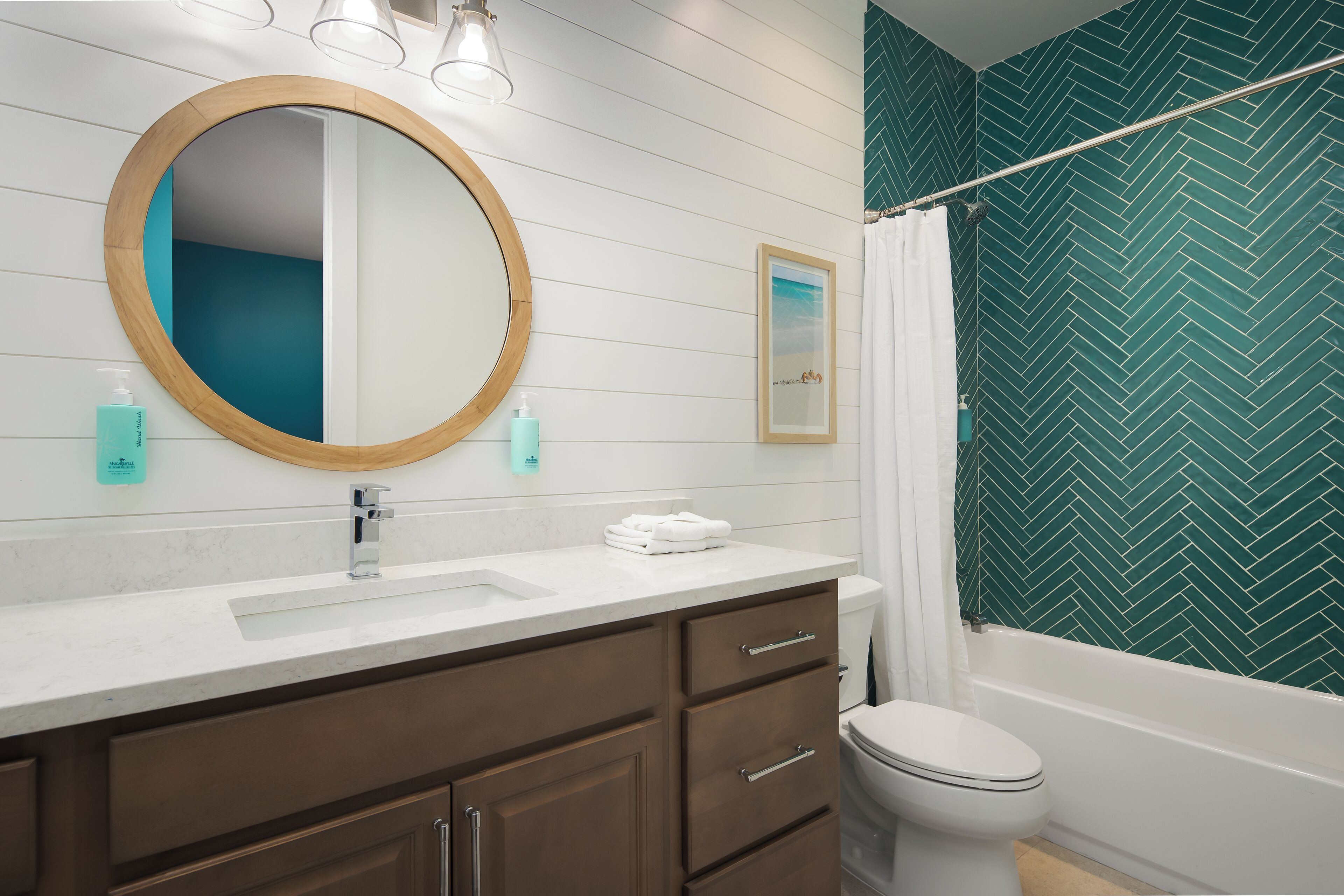 Un baño con lavamanos blanco, tocador de madera, un espejo redondo, inodoro y ducha con salpicadero de azulejos verdes en patrón de espiga.
