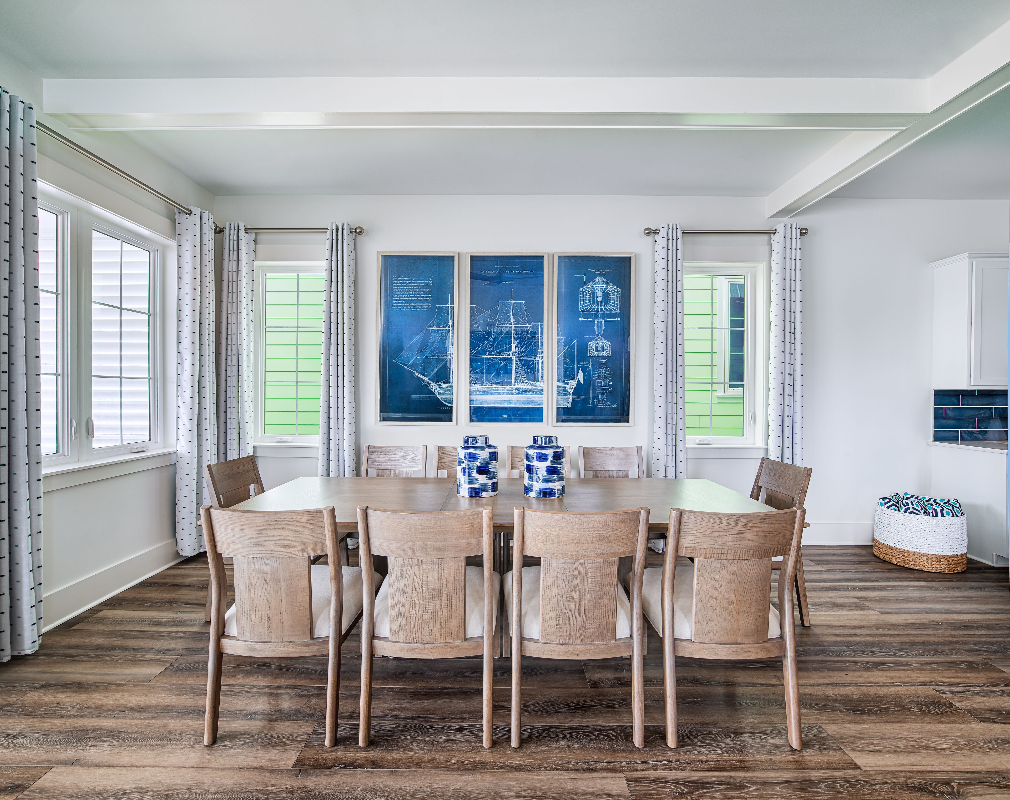 A dining area with a wooden table and chairs, large windows with curtains, and a wall-mounted ship blueprint artwork.