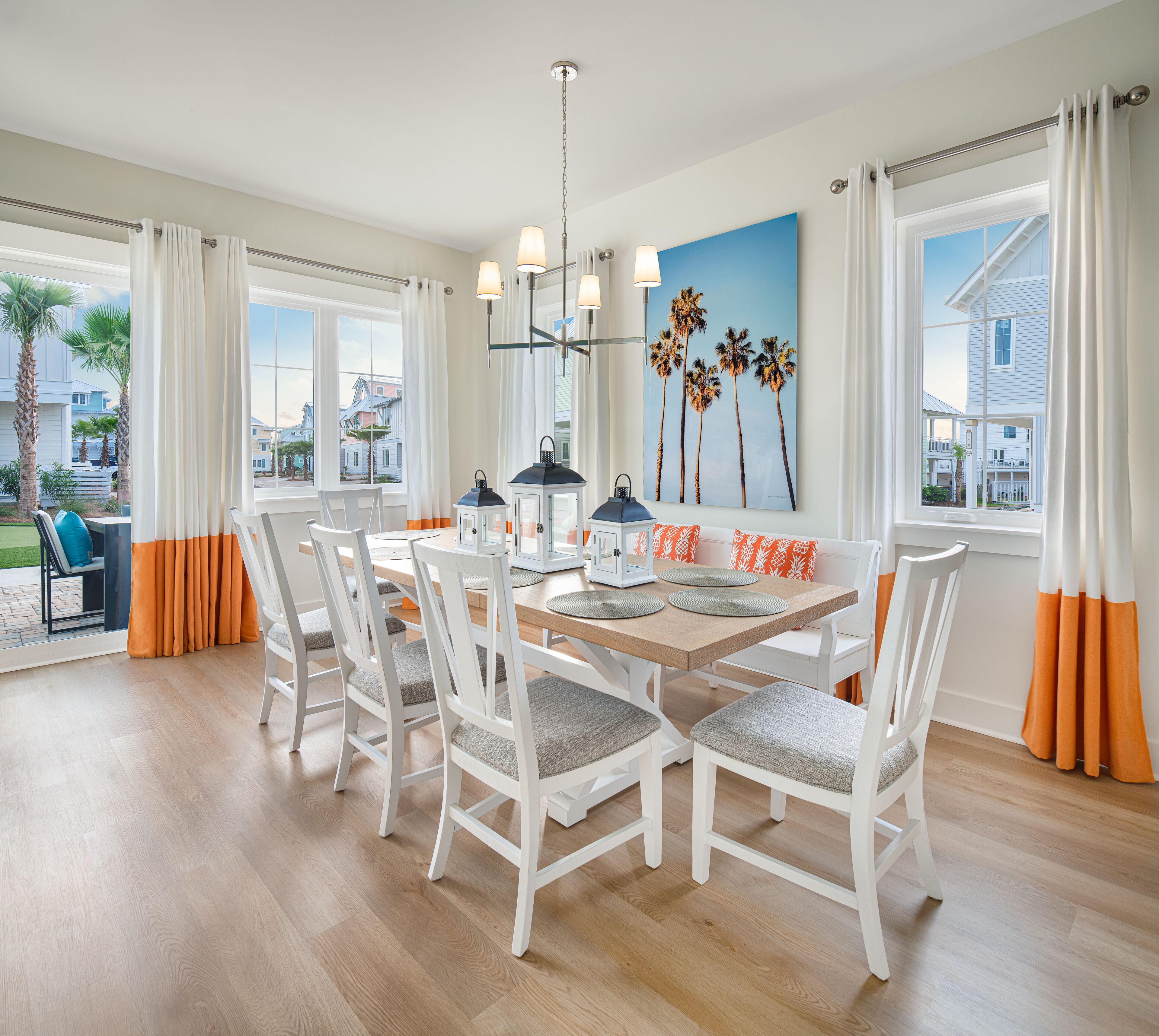 Un coin repas avec une table en bois, des chaises blanches et des coussins orange. On y trouve deux lampes fixées au mur et un tableau représentant des palmiers.