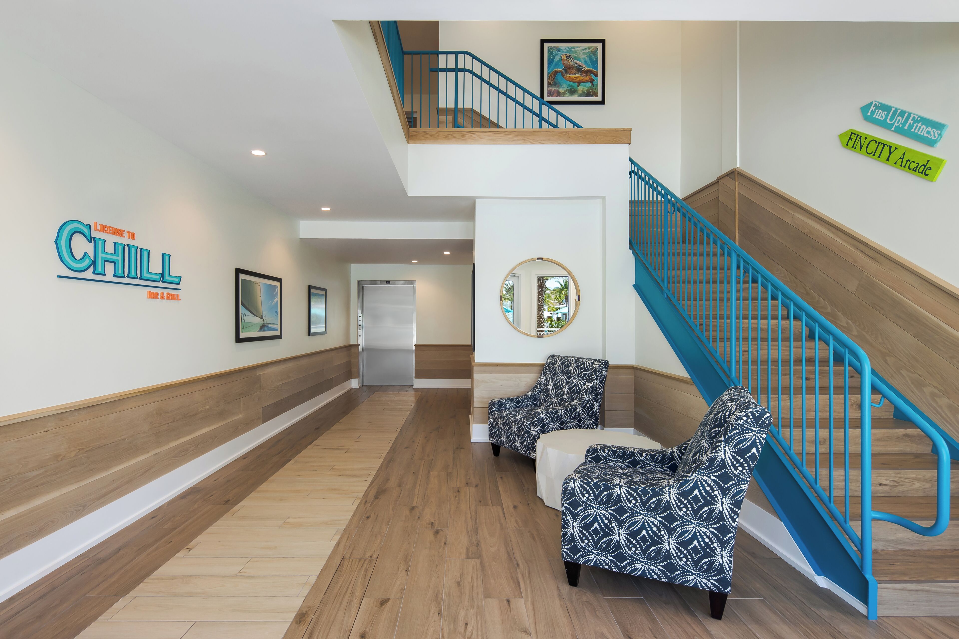 Eine Hotelhalle mit Holzboden, einer Treppe mit blauen Geländern, zwei gemusterten Sesseln und Wandbildern.