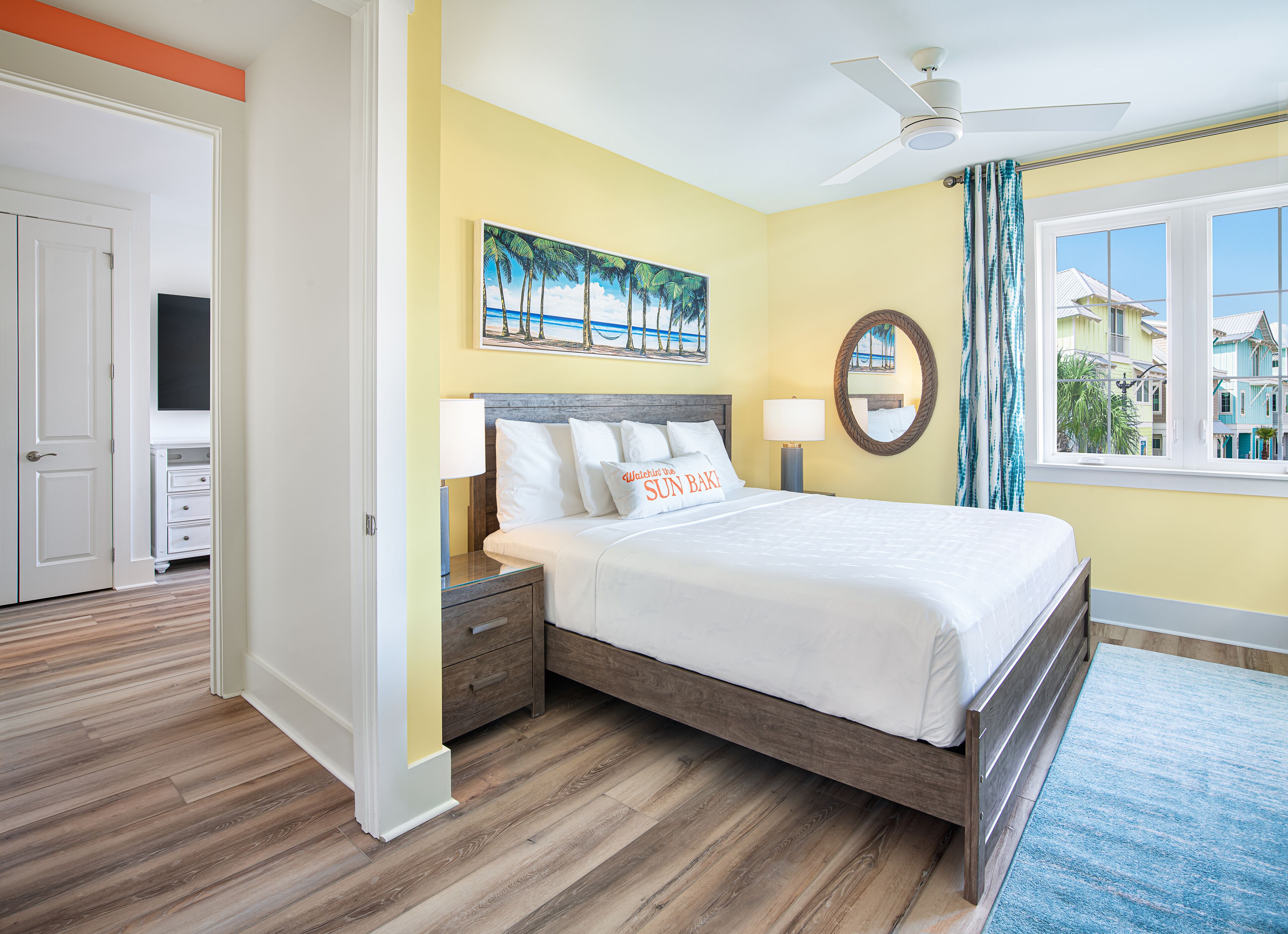 A hotel room with a bed, two bedside tables, a ceiling fan, a window with a view of buildings, and a painting on the wall.