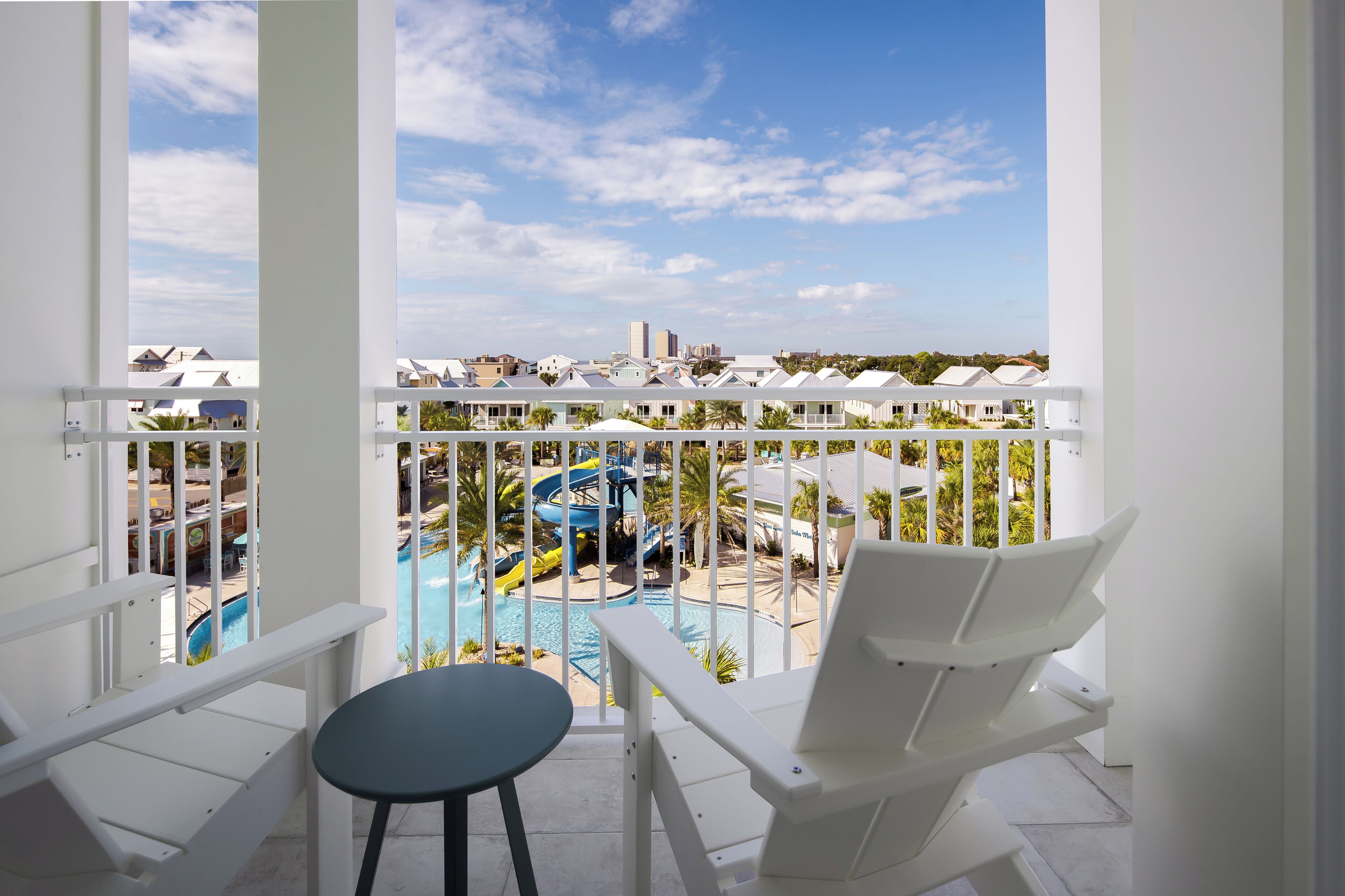 Un balcón con sillas y una mesa blancos, con vistas a una zona de piscina que incluye un tobogán y sillas de descanso.