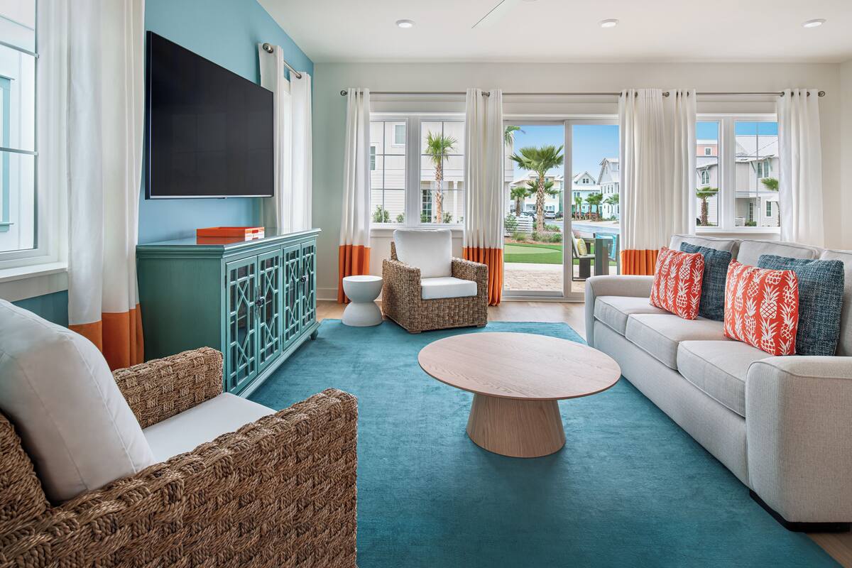 Una sala moderna con alfombra color turquesa, muebles de mimbre, un sofá blanco con cojines estampados, un televisor de pantalla plana y un ventilador de techo.