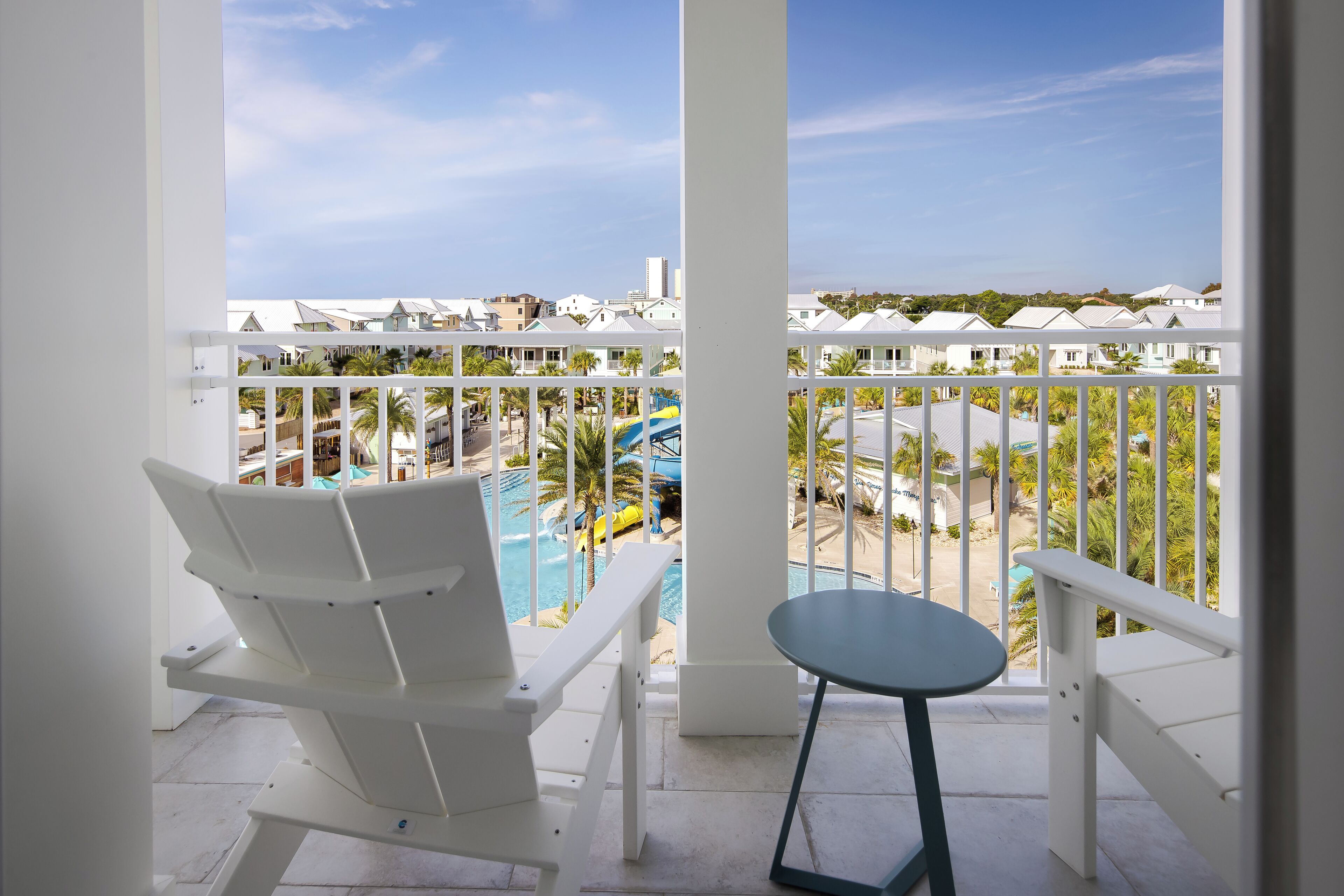 Un balcon avec des chaises blanches et une petite table, donnant sur un espace piscine équipé d’un toboggan et bordé de palmiers.