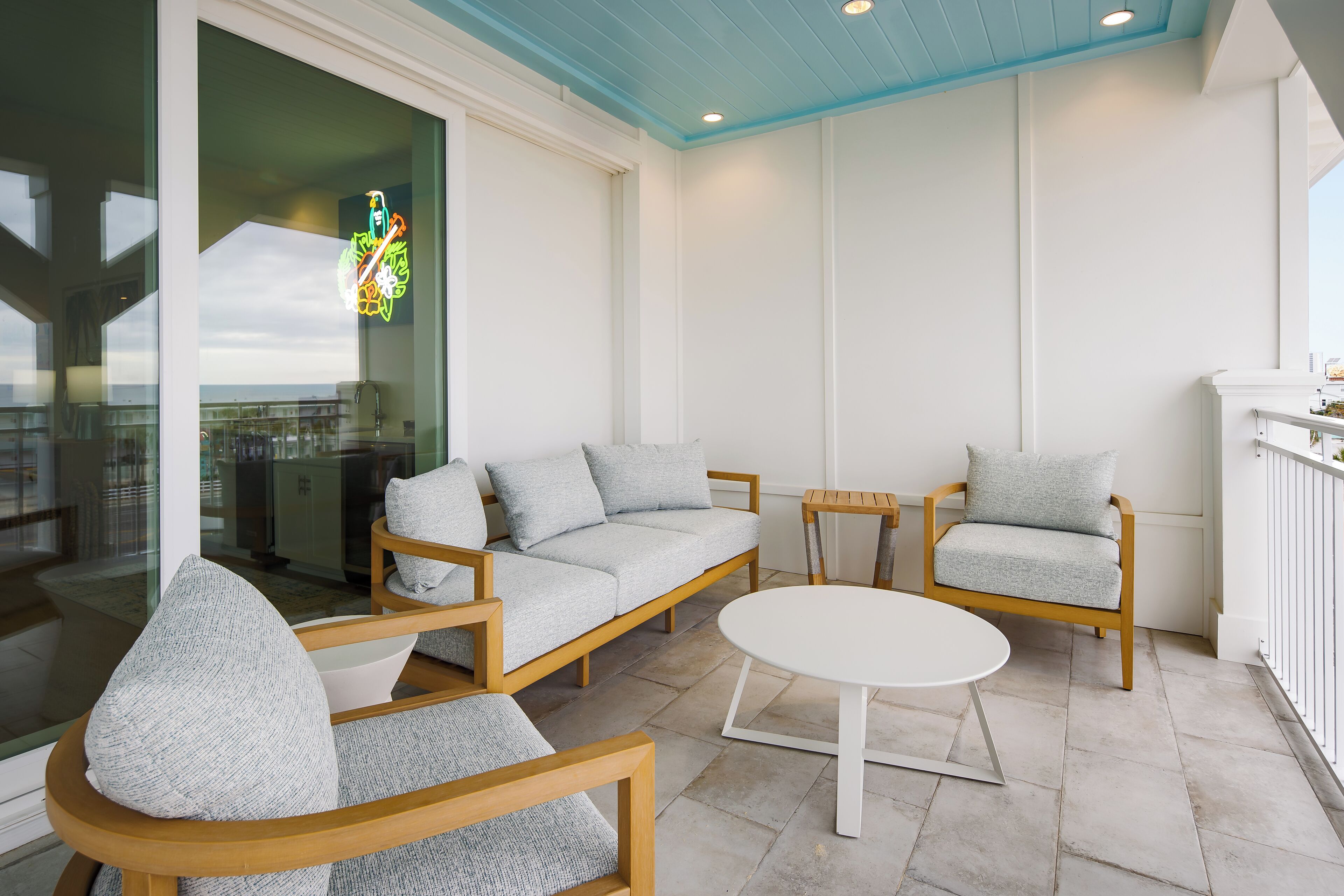 Un espace extérieur moderne avec des sièges, comprenant une table blanche et deux chaises en bois, une porte vitrée donnant sur un balcon et un panneau lumineux au-dessus de la porte.