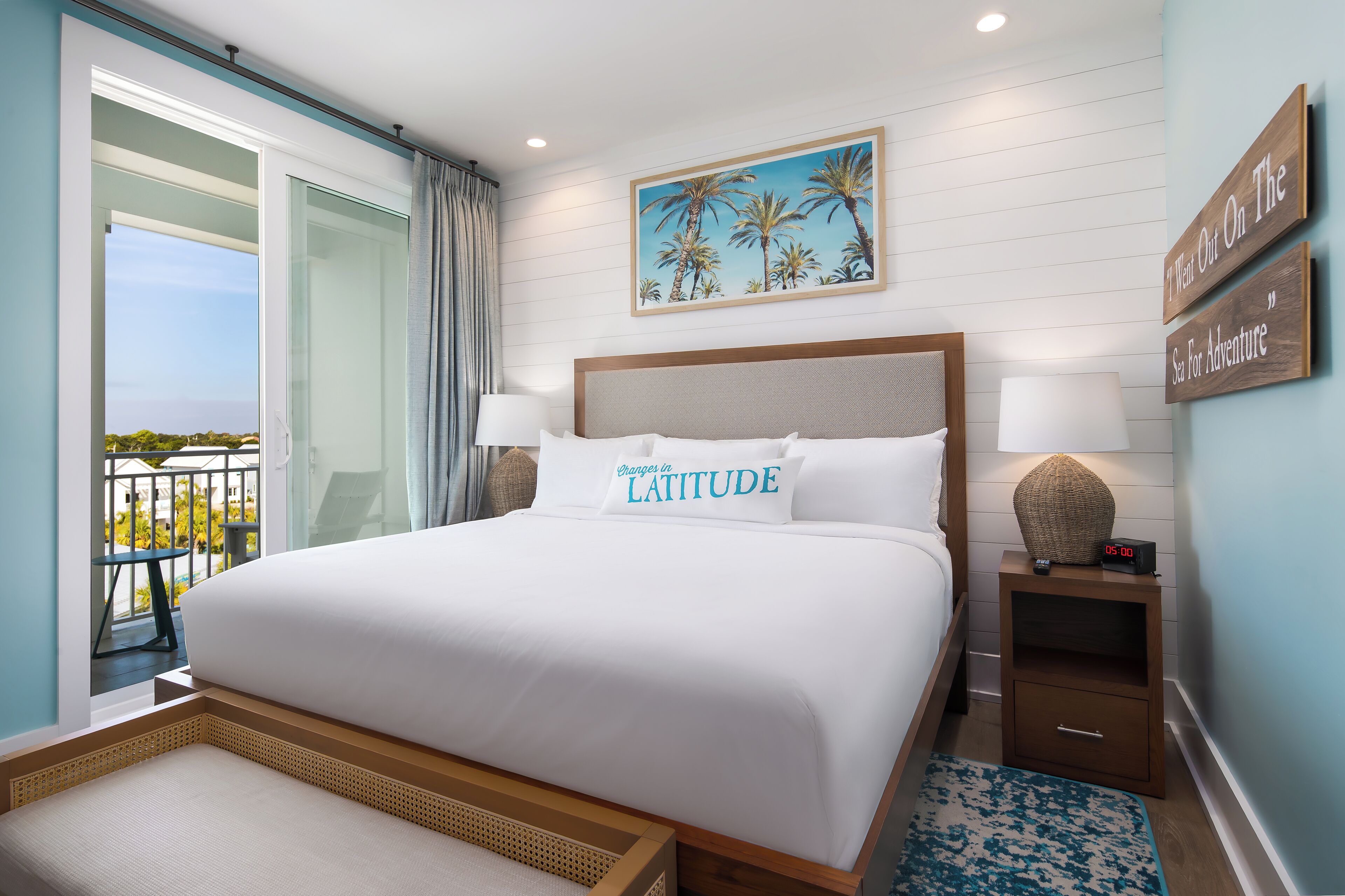 A hotel room with a large bed, a view of the outdoors, a painting of palm trees, and a sign on the wall.