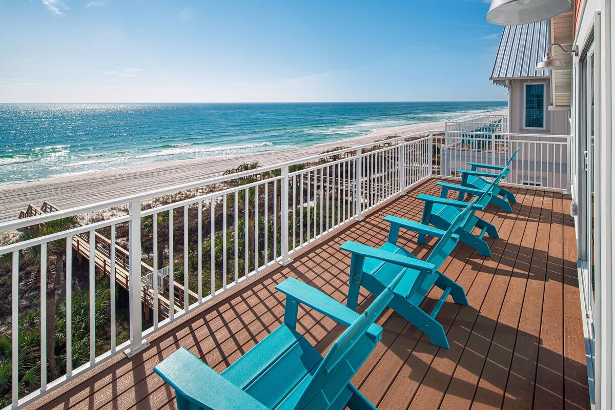 Una terraza de madera con sillas azules y barandilla blanca, con vista a una playa.