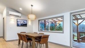 A dining area with a wooden table and chairs, a pendant light, a wall-mounted air conditioner, a framed picture, and a large window with a view of greenery. - Waterfront Cabin at White Pass, Mt. Rainier (Packwood)