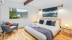 A bedroom with a large bed, a chair, a window with a view of a house and trees, and a painting of waves on the wall.