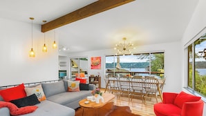 A modern living room with a grey sofa, red and black cushions, a wooden coffee table, and a red armchair. There is a dining area with wooden chairs and a chandelier. The room has large windows offering a view of the outdoors.