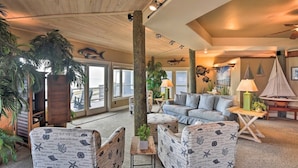 Un salon chaleureux avec un plafond en bois, un canapé, une table basse et une lampe. On y trouve des plantes et, grâce à de grandes fenêtres, une vue sur l’océan.