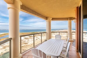 A balcony with a white table and chairs, overlooking a beach and the sea.
