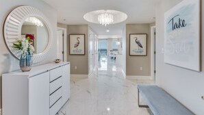A hallway with a white console, a round mirror, a chandelier, framed artwork, and a bench. - Now with lower fees! 3 Portofino 1707 (Pensacola Beach)