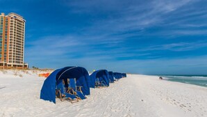 A beach with blue tents and beach chairs lined up, a tall hotel building in the background, and a clear blue sky. - Now with lower fees! 3 Portofino 1707 (Pensacola Beach)