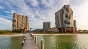 A waterfront promenade with high-rise buildings on either side, a wooden walkway, and a calm body of water. - Now with lower fees! 3 Portofino 1707 (Pensacola Beach)