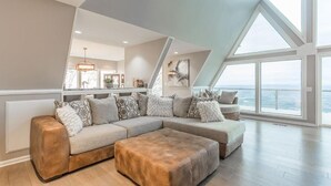 A modern living room with a sectional sofa, a brown ottoman, and a view of the sea through large windows.