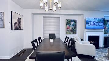 Comedor con mesa de madera oscura, sillas negras, sofá blanco, televisor y chimenea.