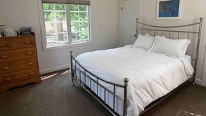 A bedroom with a metal bed frame, a wooden dresser, a window with a view of greenery, and a door.