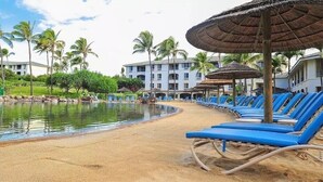 A beachside pool area with blue lounge chairs and thatched umbrellas, surrounded by palm trees and a building in the background.