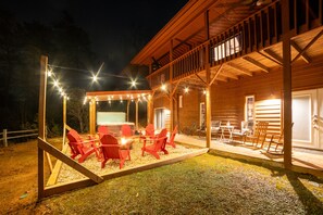 A wooden cabin with a covered outdoor seating area featuring red chairs and a table, surrounded by a wooden deck and string lights.