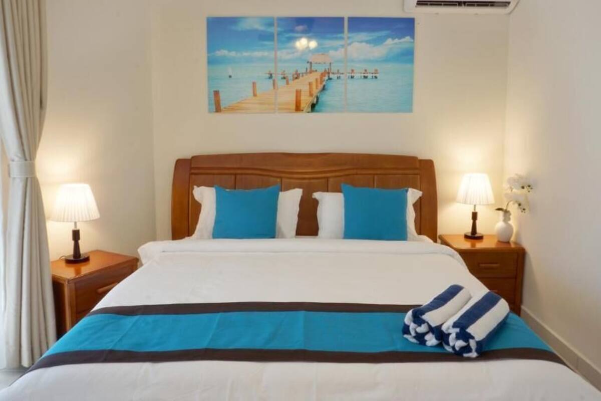 Une chambre d’hôtel avec un lit, deux tables de chevet avec des lampes, un tableau représentant une jetée accroché au mur et une fenêtre avec des rideaux.