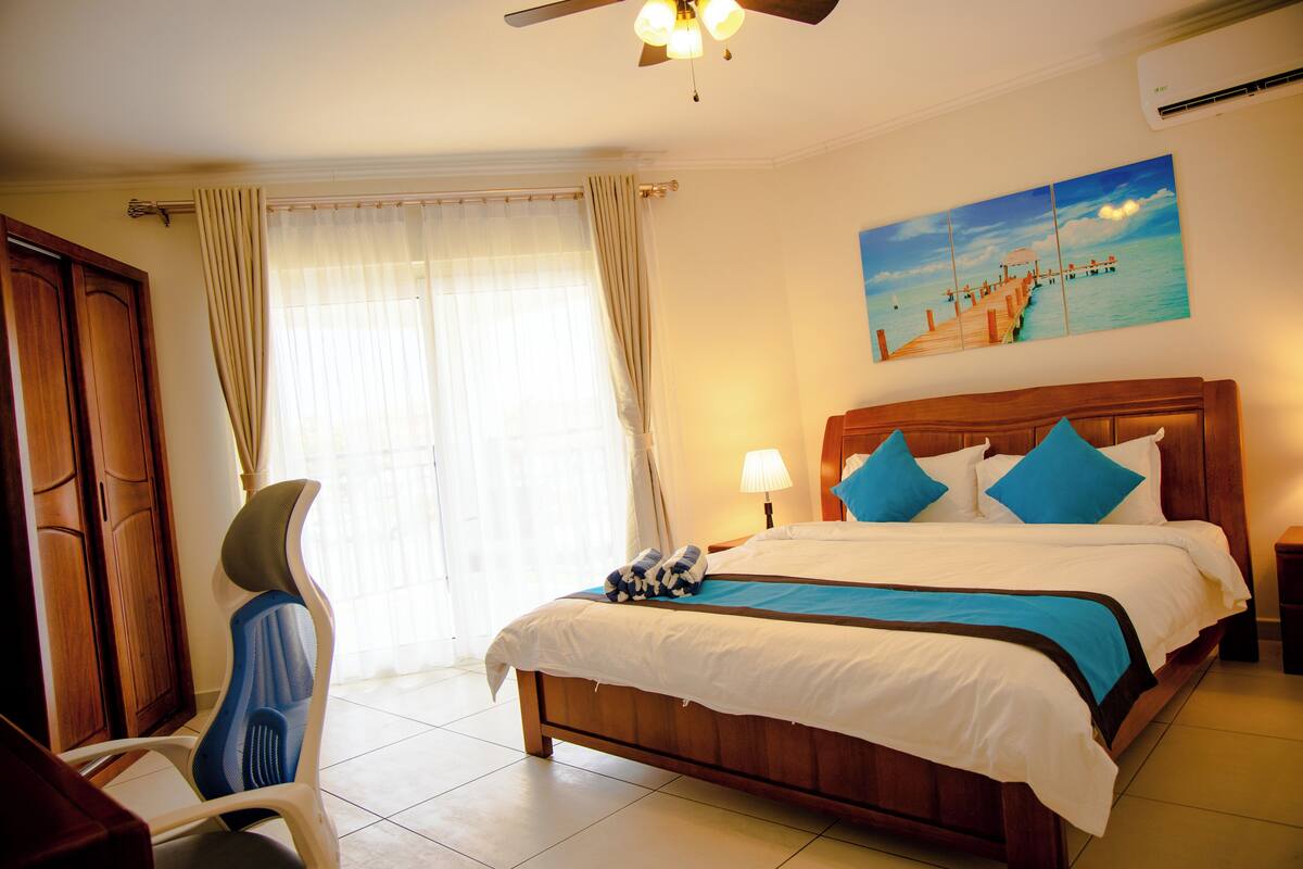 Une chambre d’hôtel avec un lit en bois, une chaise, un tableau représentant une jetée et un ventilateur de plafond.