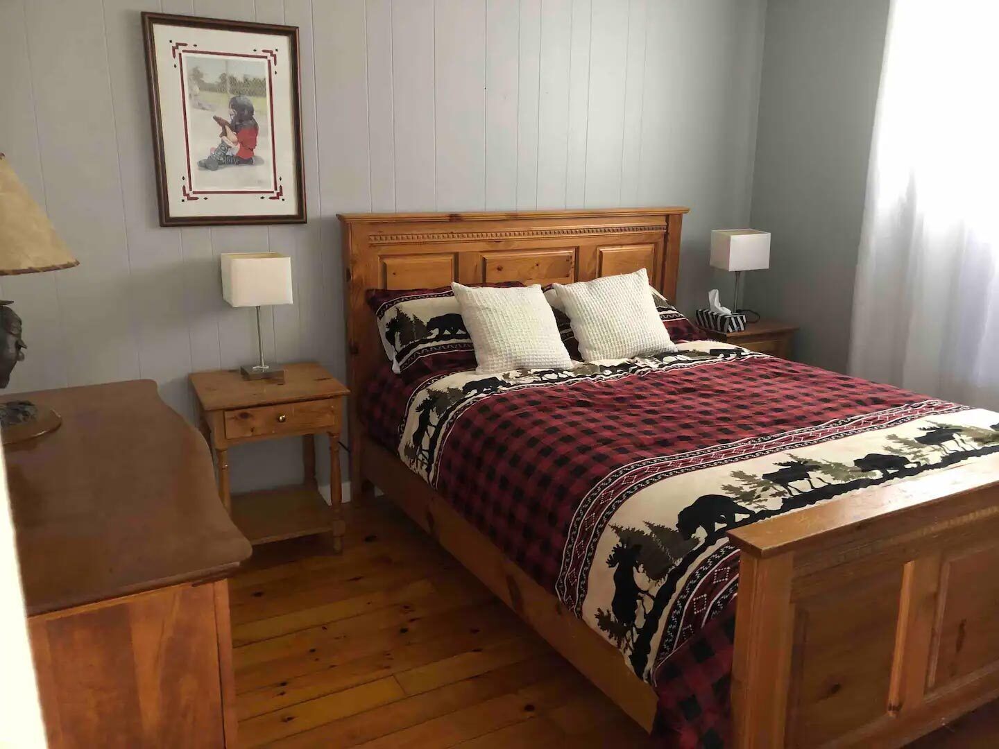 A wooden bed with a red and black checkered bedspread, two white pillows, and a headboard. There is a nightstand with a lamp and a framed picture on the wall.