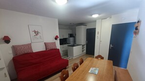 A compact living space with a red sofa, a wooden dining table, and a kitchenette in the background. - Le Corbier - Résidence Les Pistes, Appartement 6/7 Personnes au Pied des Pistes (Villarembert)