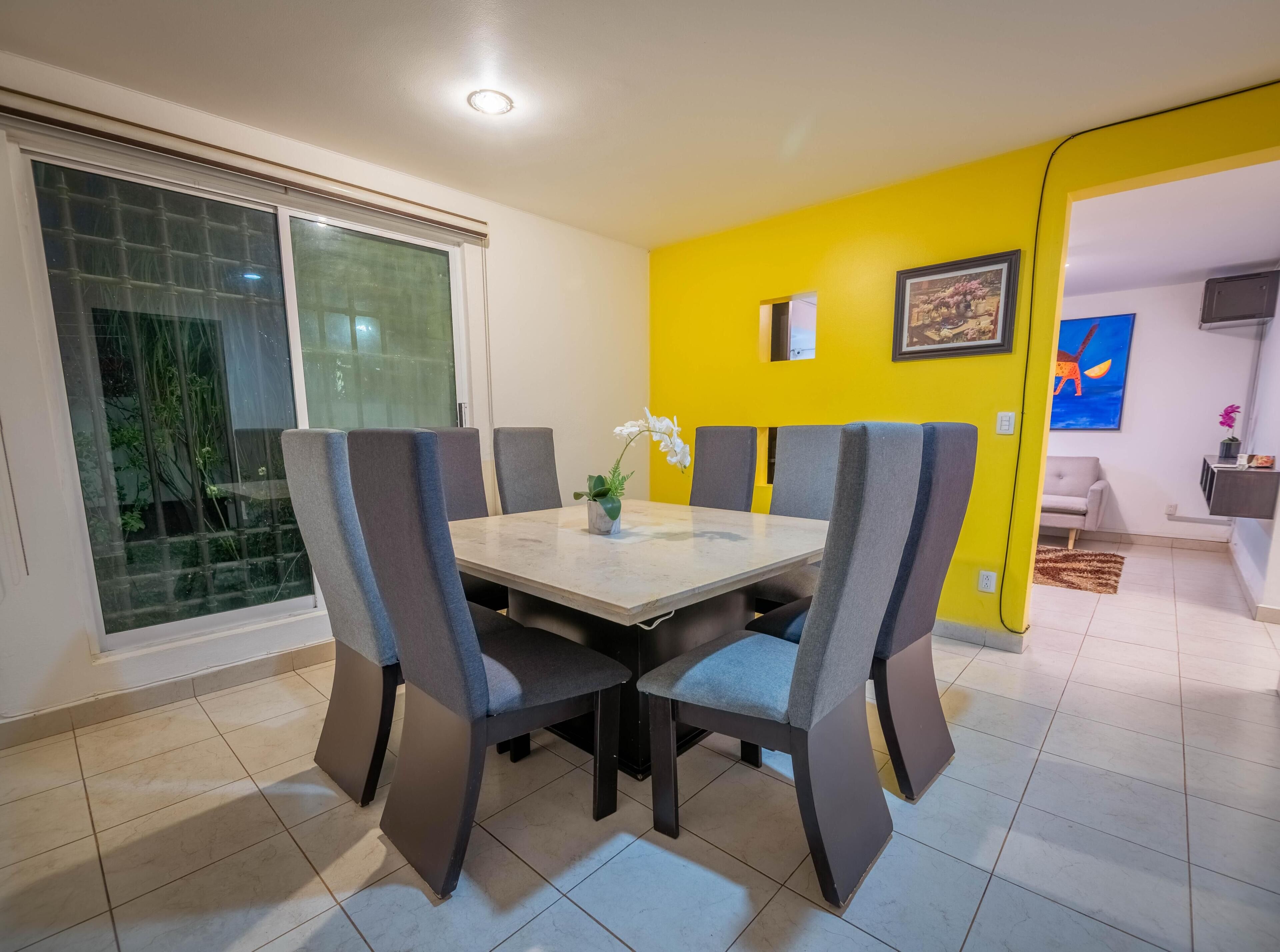 A dining area with a table and chairs, a window with a view, and a bright yellow wall.