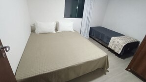 Une petite chambre propre, avec un lit simple, une fenêtre avec des rideaux et une table de chevet sur laquelle est posée une couvre-lit à motifs.